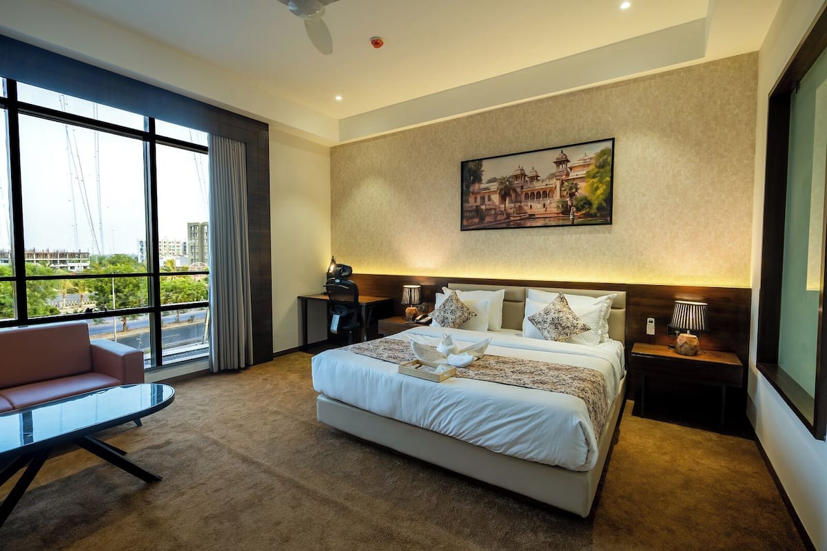 Ruime hotelkamer met een groot bed, een bureau en stadszicht door raamhoog plafond.
