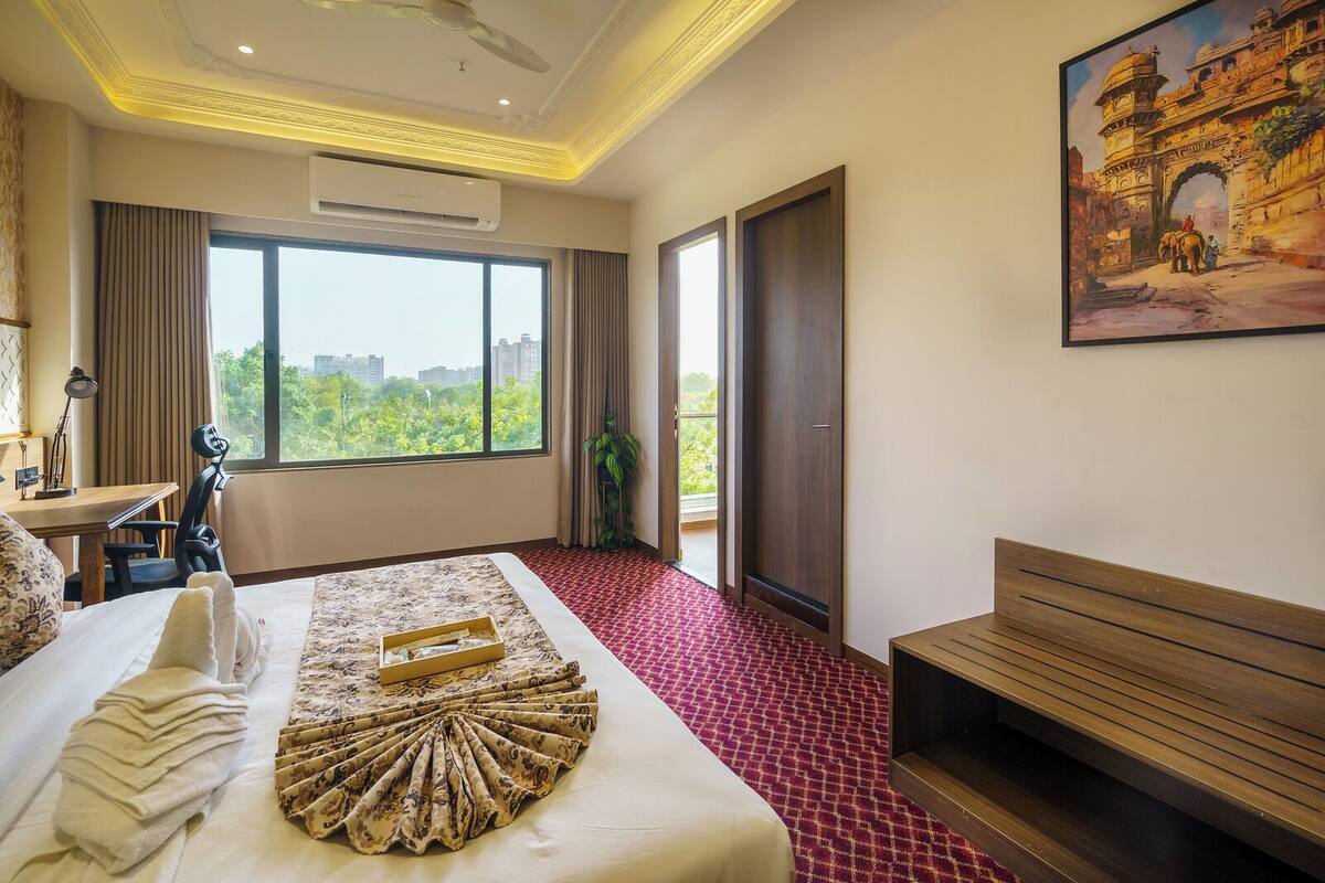 Een hotelkamer met een bed, een bureau, een stoel, een bank, een groot raam met uitzicht op de stad en een schilderij aan de muur.