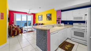 A bright kitchen with white cabinets, a microwave, and an oven. There is a colorful sofa and a dining area with a view of the sky.