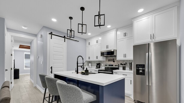 A modern kitchen with white cabinets, a stainless steel refrigerator, a central island with bar stools, and pendant lighting.