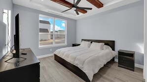 A bedroom with a bed, a nightstand, a TV on a desk, a ceiling fan, and a window with a view of houses.