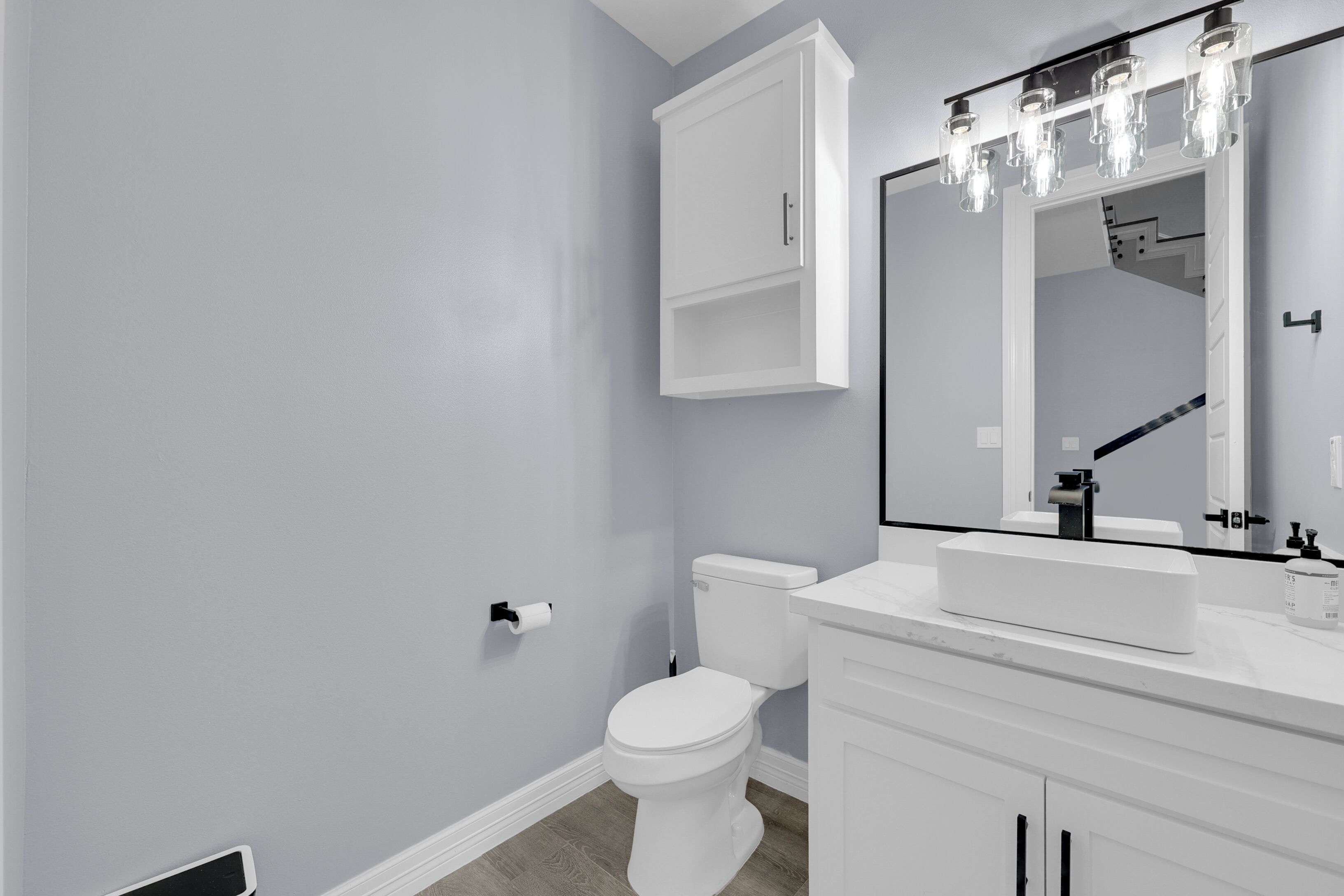 Une salle de bain avec des toilettes blanches, un lavabo blanc et un miroir au-dessus du lavabo. Il y a un meuble au-dessus des toilettes et un luminaire au-dessus du miroir.