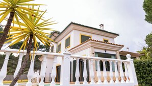 A two-story building with a white balcony railing, a palm tree in the foreground, and a clear sky.