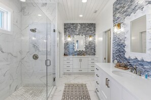 Une salle de bain dotée d’une douche à parois de verre, d’un sol carrelé en marbre, d’un pare-chocs décoratif et d’un meuble-lavabo blanc avec des poignées dorées.