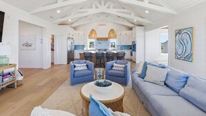 A spacious living area with a kitchen in the background, featuring white cabinetry, blue bar stools, and a large window.