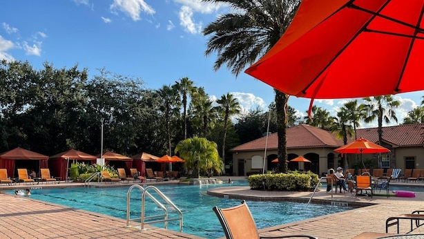 Un área de piscina con sillones de descanso y sombrillas, rodeada de palmeras y un edificio con tejado de tejas.