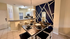 Una cocina moderna con armarios blancos, microondas y estufa. Hay una zona de comedor con una mesa de madera, cuatro sillas y un espejo decorativo en la pared.