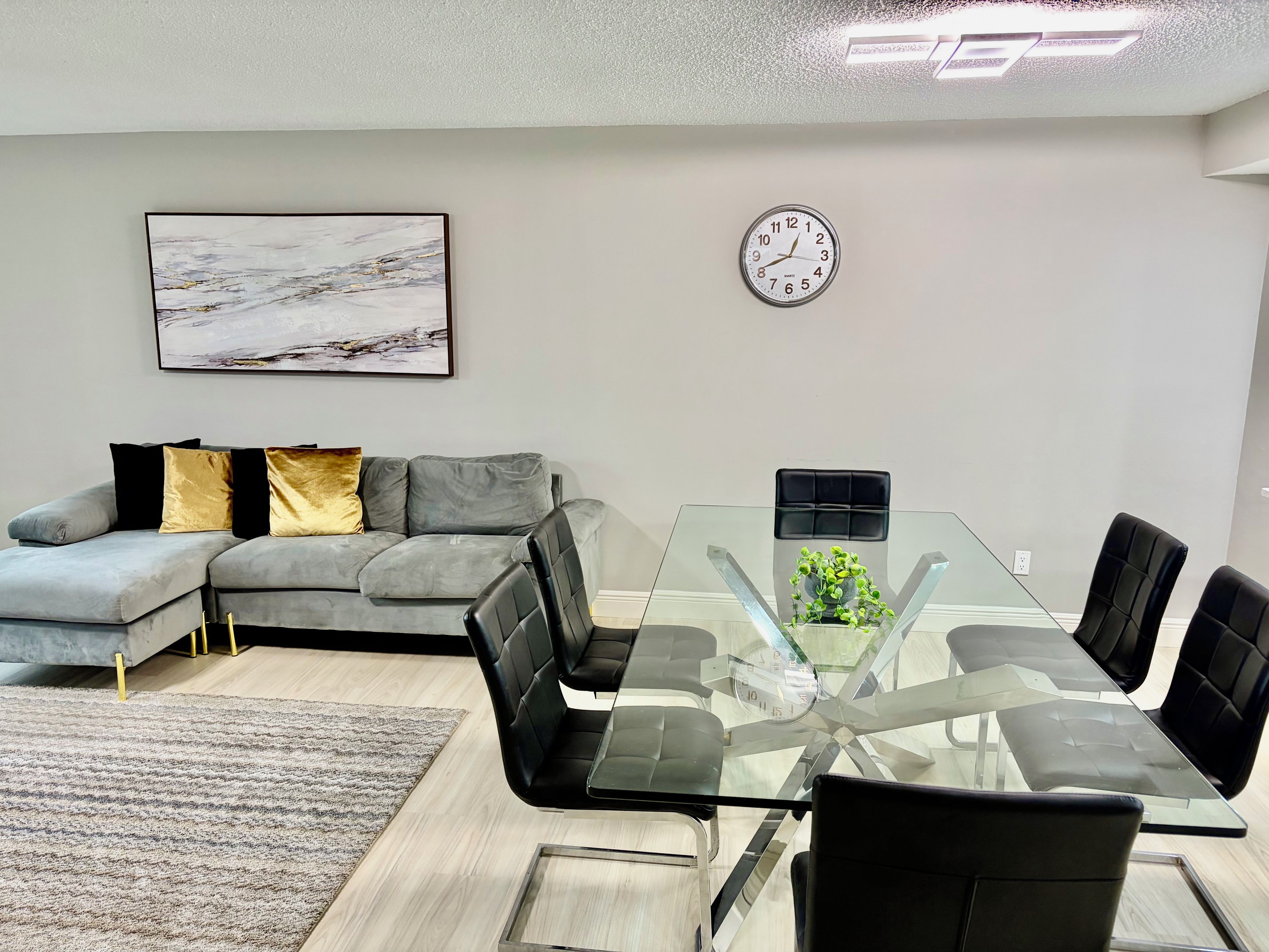 Un salon moderne avec une table à plateau en verre, des fauteuils en cuir noir, un canapé gris agrémenté de coussins dorés et noirs, une horloge au mur et un tableau abstrait encadré.
