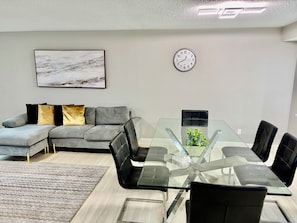 Ein modernes Wohnzimmer mit einem Glastisch, schwarzen Ledersesseln, einem grauen Sofa mit goldenen und schwarzen Kissen, einer Uhr an der Wand und einem gerahmten abstrakten Gemälde.