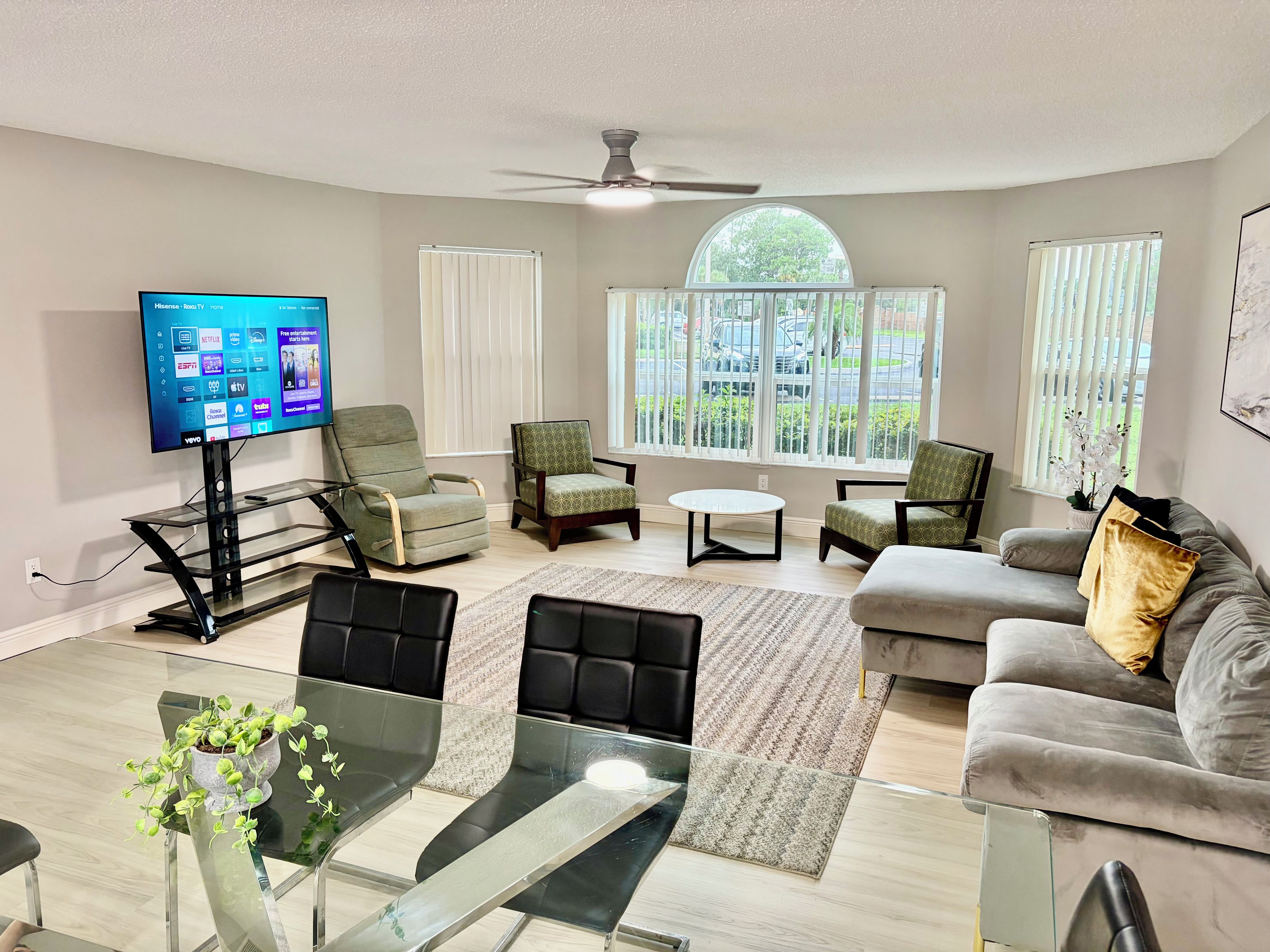 A modern living room with a flat-screen TV, multiple seating options, a glass coffee table, and a view of the outdoors through large windows.