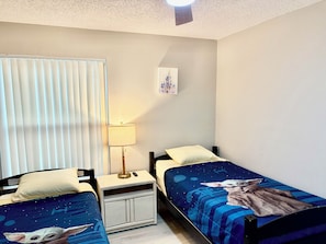 Un dormitorio con dos camas individuales, una mesita de noche con lámpara, una ventana con persianas y un cuadro colgado en la pared.