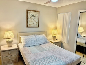 Ein Schlafzimmer mit einem Bett, zwei Nachttischen mit Lampen, einem Schminktisch, einem Deckenventilator, einem Fenster mit Jalousien und einem gerahmten Bild an der Wand.