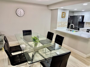 A modern kitchen with a glass dining table, black chairs, a clock, and a refrigerator.