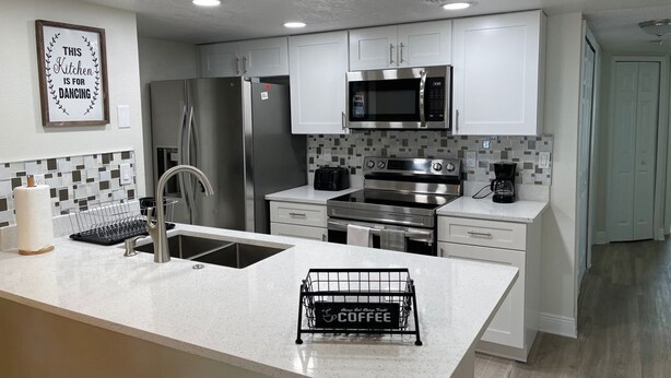 A modern kitchen with white cabinets, stainless steel appliances, a tiled backsplash, and a kitchen island with a coffee station. - Grand Disney Somerset 3BR Condo Near Parks (Kissimmee)
