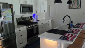A modern kitchen with white cabinets, stainless steel appliances, a central island with stools, pendant lighting, and a wall clock.