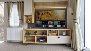 A modern living room with a large flat-screen TV mounted on a wooden wall, a built-in shelf with decorative items, a radiator, and a window with curtains.