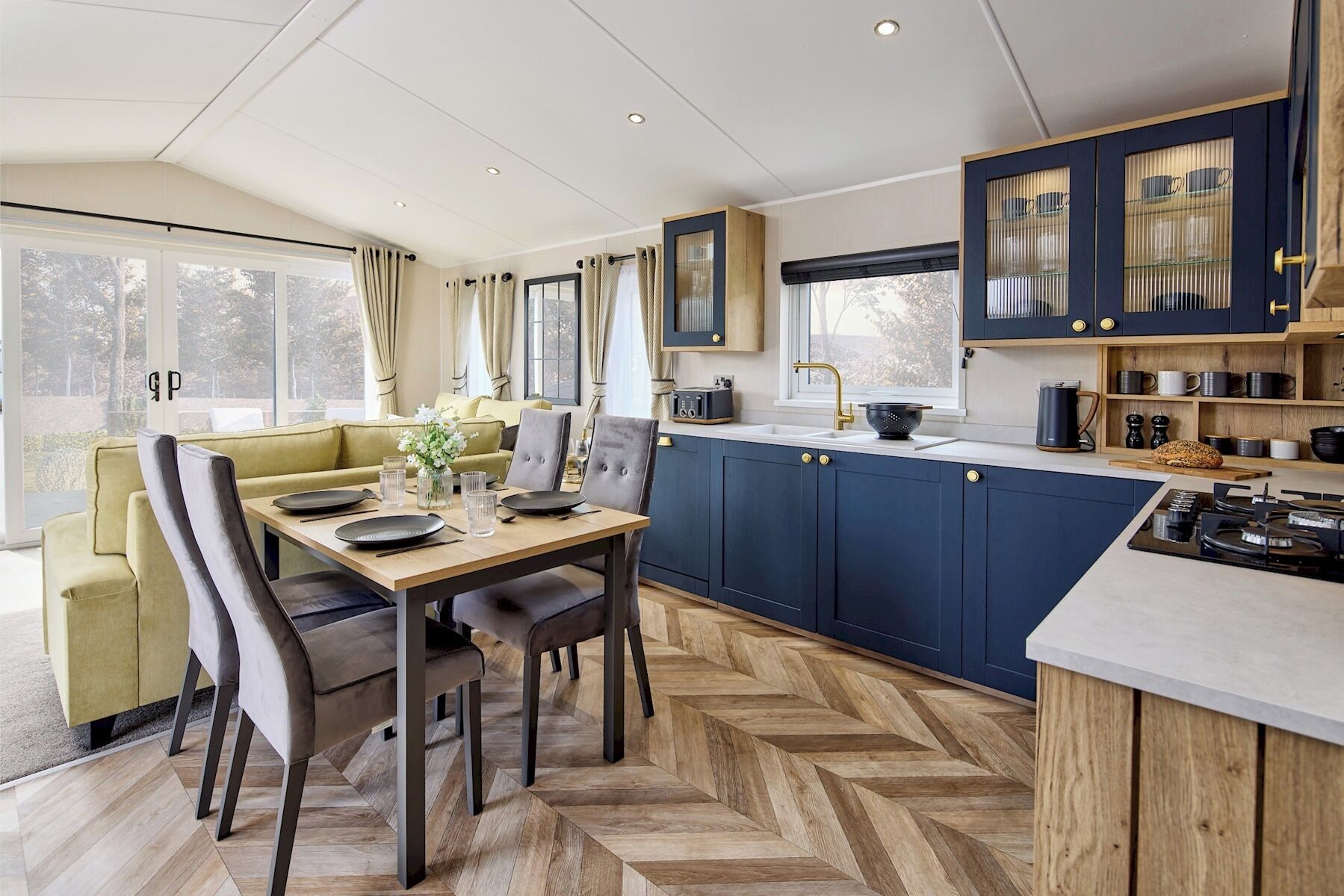 Eine moderne Küche mit dunkelblauen Schränken, einem Fischgrätparkett und einem Essbereich mit einem grünen Sofa und einem Holztisch.