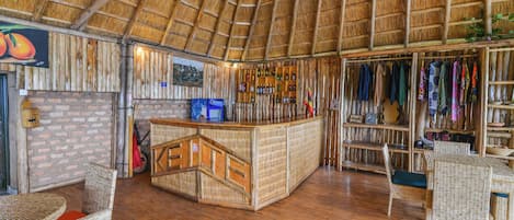 A rustic bar area with a thatched roof, wooden furniture, and a variety of bottles on shelves.