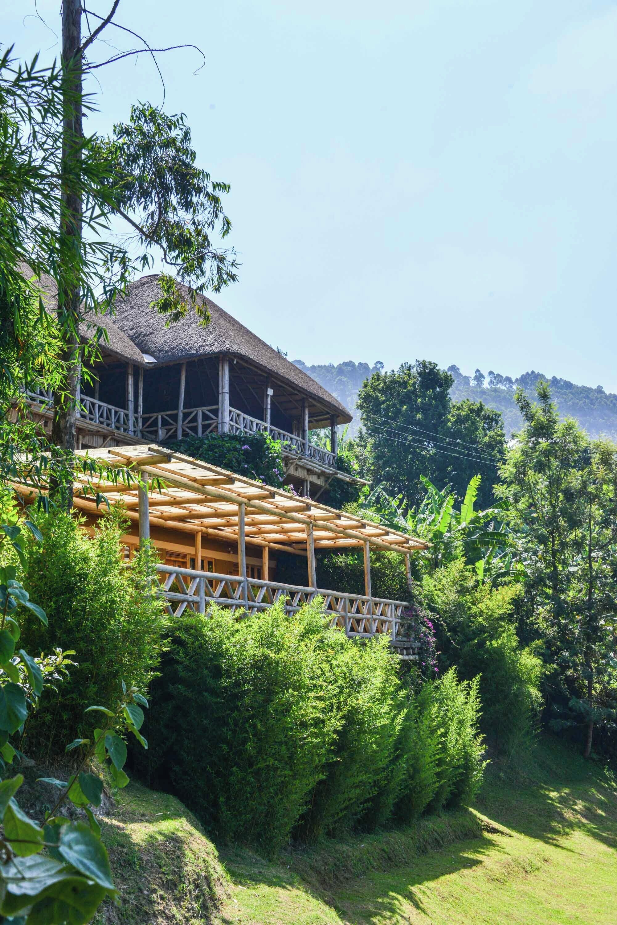 A traditional wooden building with a thatched roof, surrounded by lush greenery and a clear view of the mountains in the background.