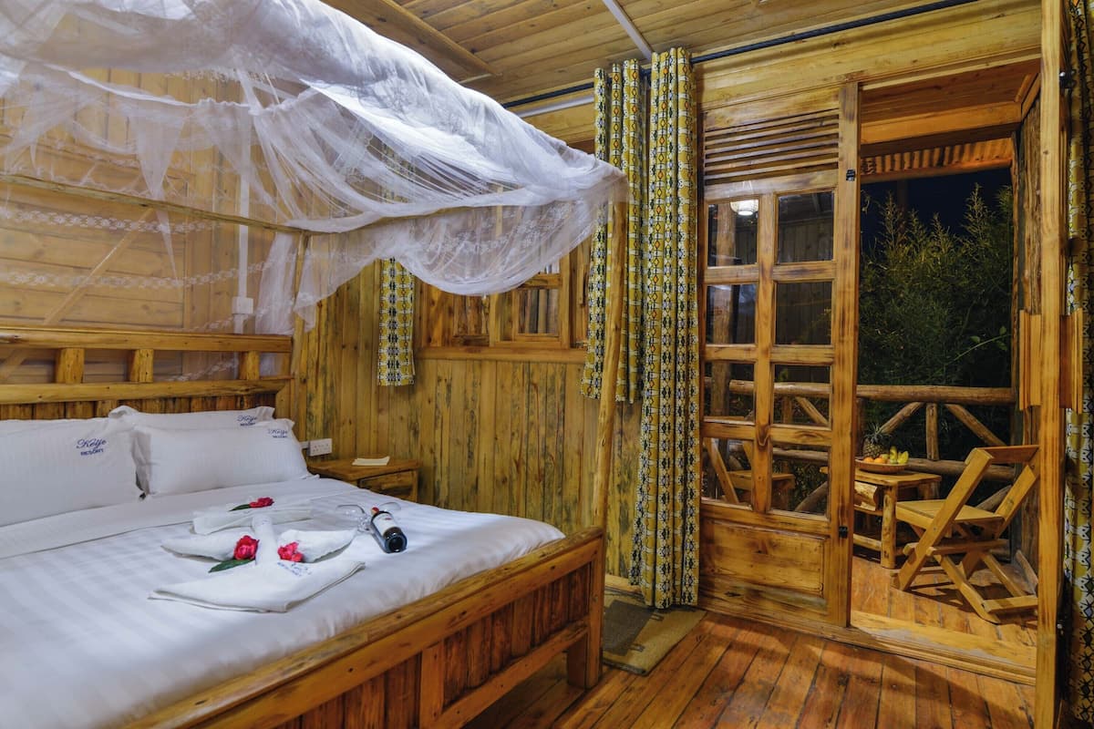 Ein Zimmer in einer Holzhütte mit einem Bett, einer Holzbank, einem Tisch mit Früchten und einem Fenster mit Vorhängen.