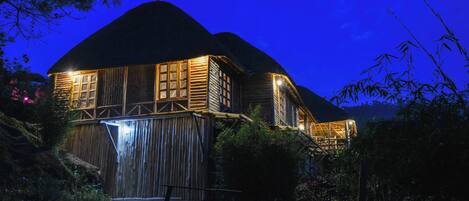 A rustic wooden house with a thatched roof, surrounded by lush greenery and bamboo.