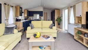 A spacious living area with a yellow sofa, a coffee table with magazines and fruit, and a kitchen in the background.