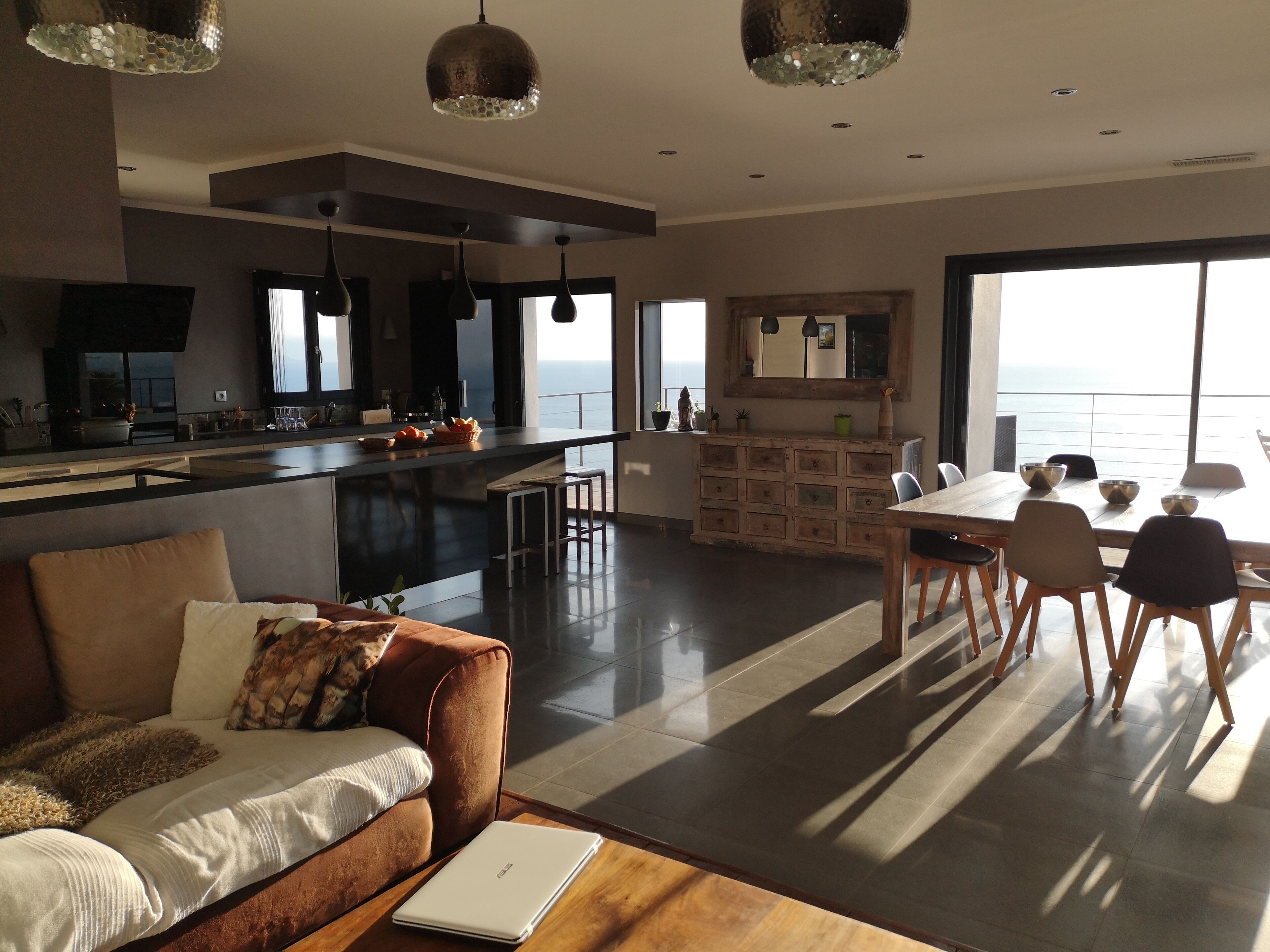 Un salon moderne comprenant un canapé, une table basse et un coin repas avec une table et des chaises. Il y a également une cuisine avec un comptoir et une vue sur la mer.