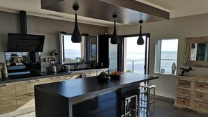 Une cuisine moderne dotée d’un grand îlot central, de luminaires suspendus et offrant une vue sur l’océan.