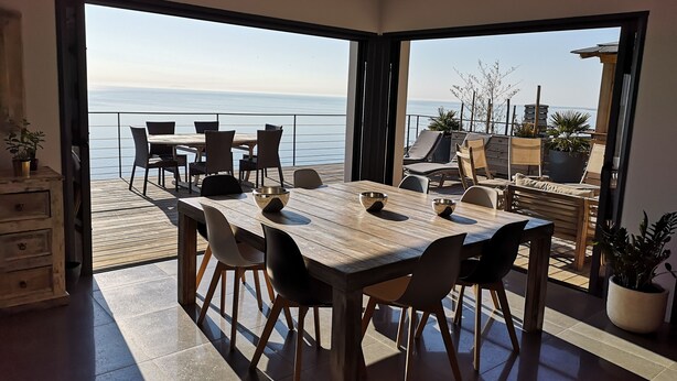 Un espace repas offrant une vue sur la mer, avec une table et des chaises en bois, ainsi qu’un balcon aménagé avec des sièges.
