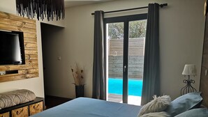 Une chambre à coucher avec un lit, un téléviseur fixé au mur en bois, une vue sur une piscine à travers une porte coulissante et une lampe sur une table de chevet.