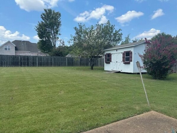 A small white shed with a door and windows, surrounded by a grassy yard and a wooden fence.