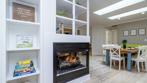 A modern living room with a fireplace, white shelving with books and decorative items, a dining area with a wooden table and chairs, and a bright ceiling with skylights. - (3 Nt Summer Min!) Union Pier Condo With Heated Pool! July 04-10 OPEN! (Union Pier)