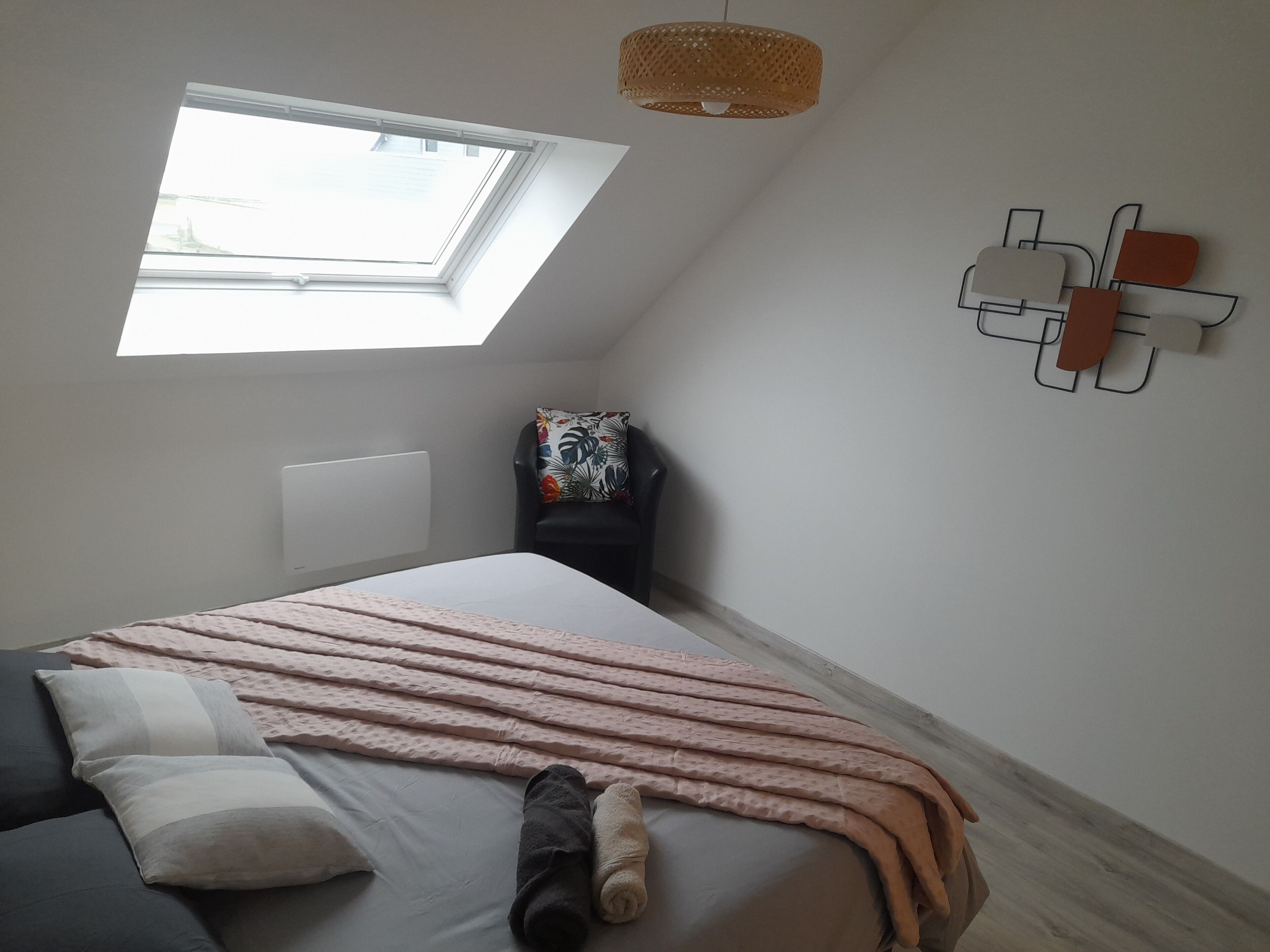 Ein Schlafzimmer mit Dachfenster, einem Bett mit Kissen und Decke, einem Sessel mit Blumenkissen und einer wandmontierten Lampe.