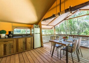 Une terrasse en bois avec une table et des chaises, un réfrigérateur, une machine à café et une fenêtre donnant sur la verdure.