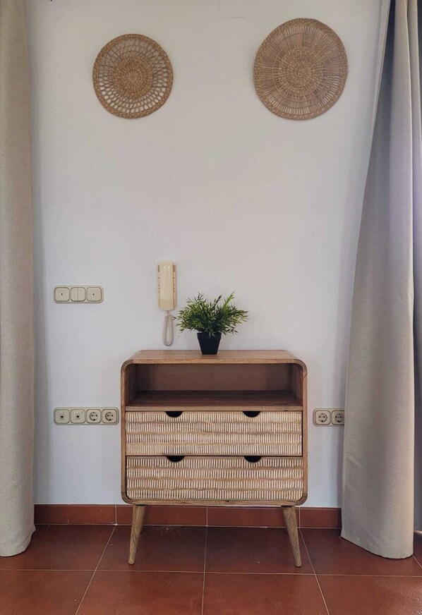 Quarto com mesa de consola de madeira, uma planta em vaso, duas tapeçarias tecidas, telefone e tomadas elétricas.