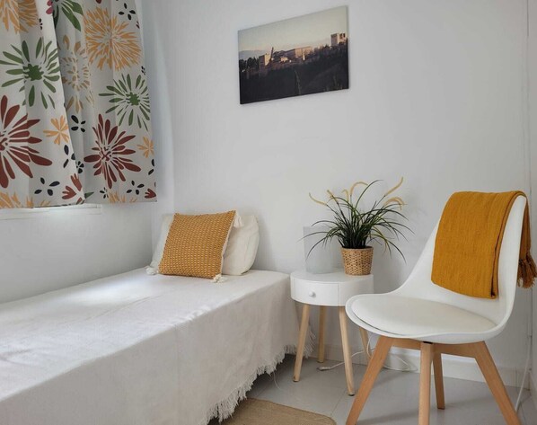Ett sovrum med en säng, en stol, ett litet bord, en krukväxt och en målning med ett stadsmotiv på väggen.