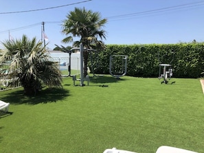 A well-maintained lawn with exercise equipment, palm trees, and a hedge.