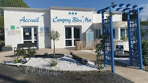 Un edificio con letreros que dicen "Accueil" y "Camping Bleu Noir", una banca y una estructura de pérgola azul.