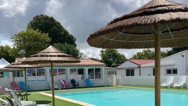 Una piscina con sillas de descanso blancas y sombrillas de paja, rodeada por un edificio blanco con techo rojo.