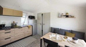 A modern kitchen with wooden cabinets, a gas stove, and a microwave. There is a dining area with a table, chairs, and a bowl of fruit. - Camping 3 étoiles - Piscine  - ccbahcd (Saint-Georges-de-Didonne)