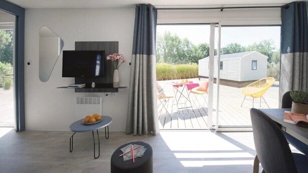 Habitación moderna con televisor de pantalla plana, una mesita con fruta, una silla y un balcón con mesa y sillas.