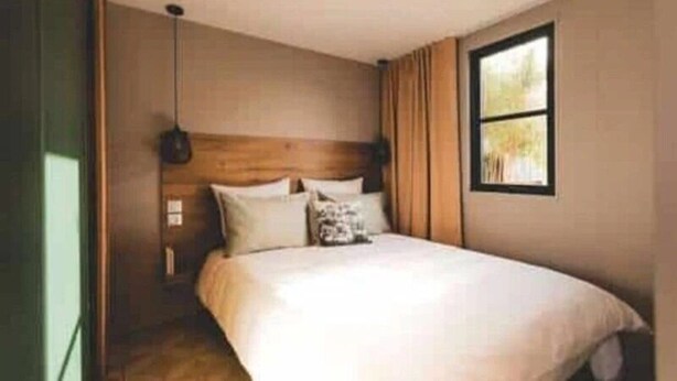 Ein Schlafzimmer mit einem hölzernen Kopfteil, einem Bett mit weißer Bettwäsche, einer Pendelleuchte und einem Fenster mit Vorhängen.
