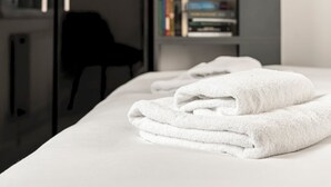 Un lit bien fait, avec des serviettes blanches pliées, une chaise noire et une bibliothèque remplie de livres, dans une chambre au style moderne.