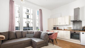 Un salon moderne comprenant un canapé d’angle, un coin repas et une cuisine avec des meubles blancs et des appareils électroménagers en acier inoxydable.