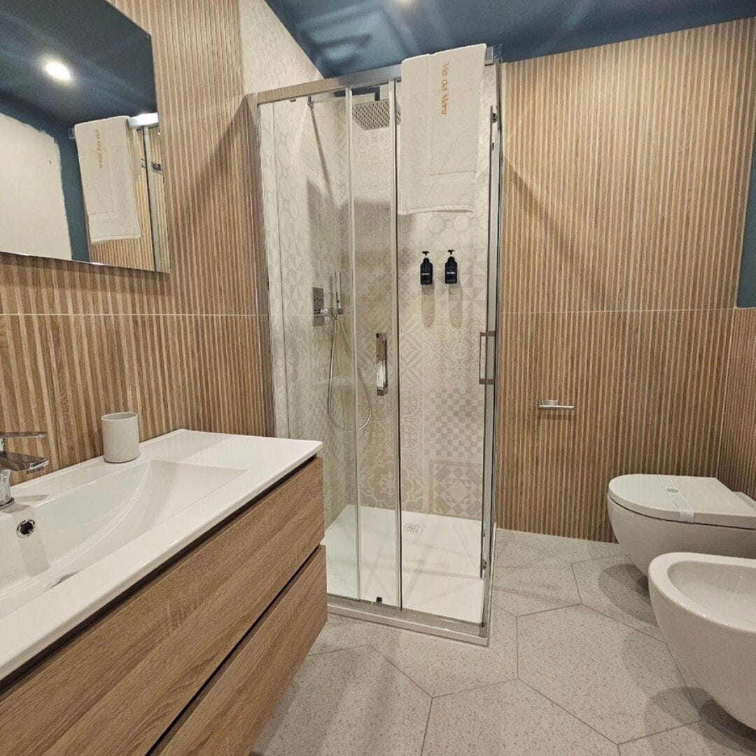 Een moderne badkamer met een glazen douchecabine, een houten wastafelmeubel, een toilet en een bidet.