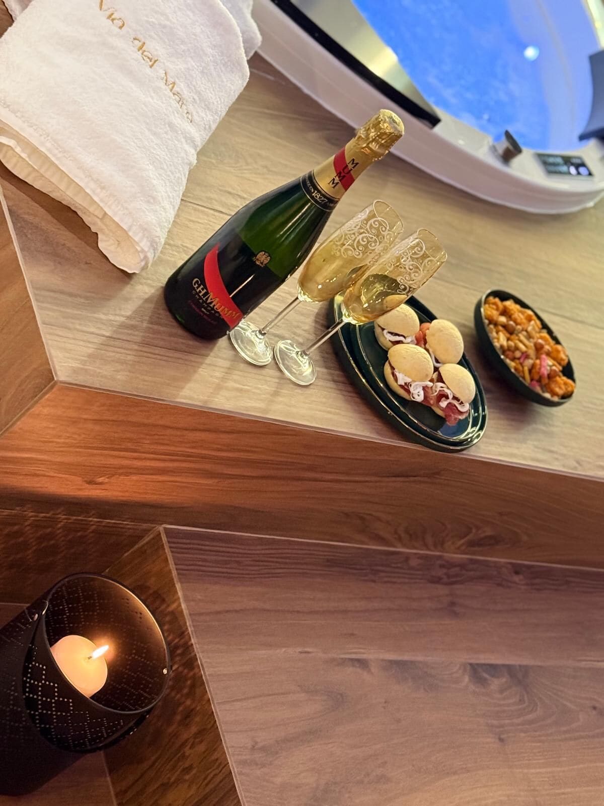 Et træbord med en champagneflaske, to glas, en tallerken med småkager, et stearinlys og et håndklæde.