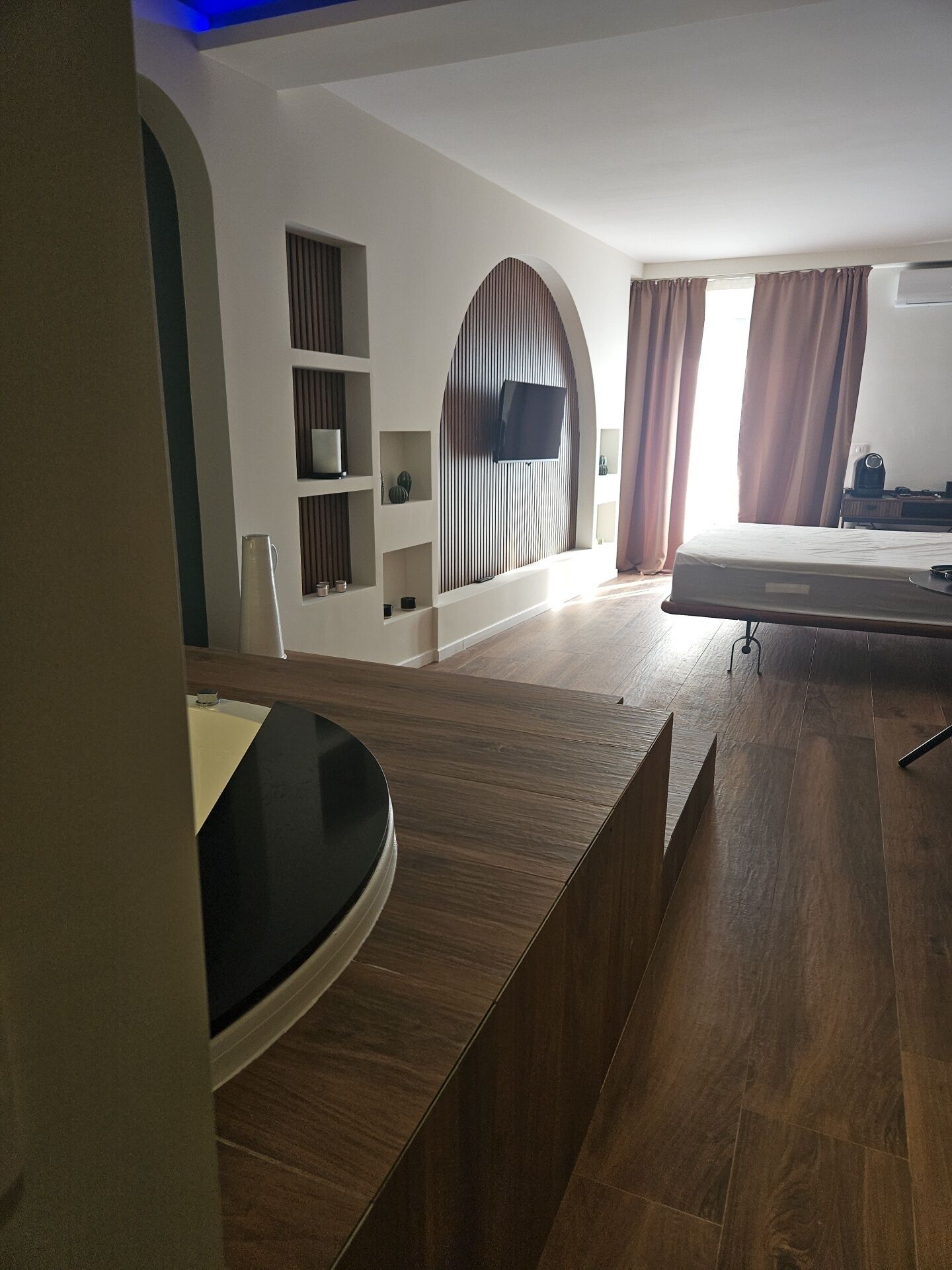 Una habitación de hotel moderna con un mostrador de recepción de madera, un televisor de pantalla plana montado en la pared, una cama con colchón y almohadas, y una mesita con un hervidor.