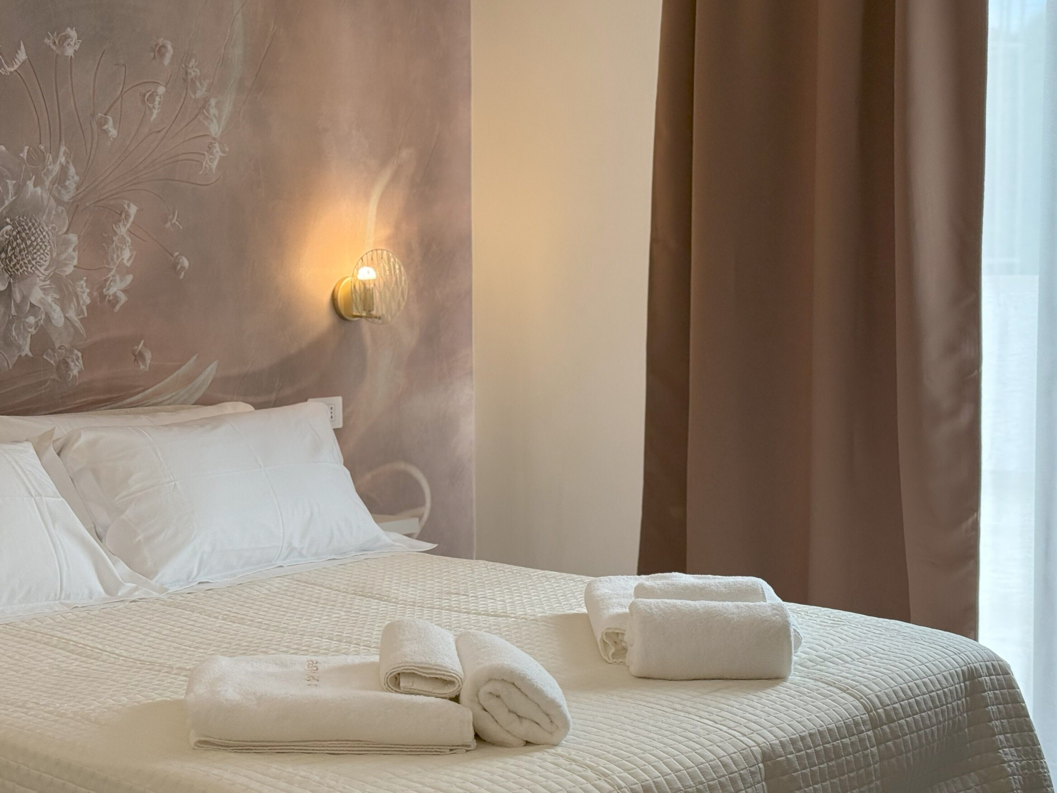 Una cama bien hecha con sábanas blancas y toallas enrolladas, una lámpara de pared y una ventana con cortinas.