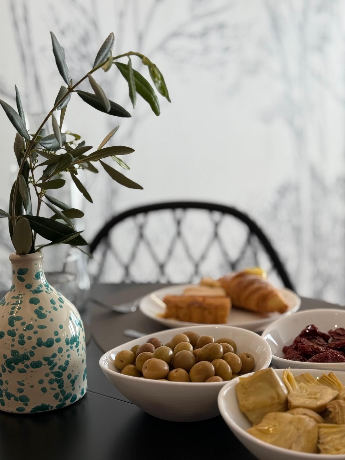 Une table à manger ornée d’un vase de feuilles vertes, d’un bol d’olives, d’une assiette de croissants et d’un bol de cœurs d’artichauts.