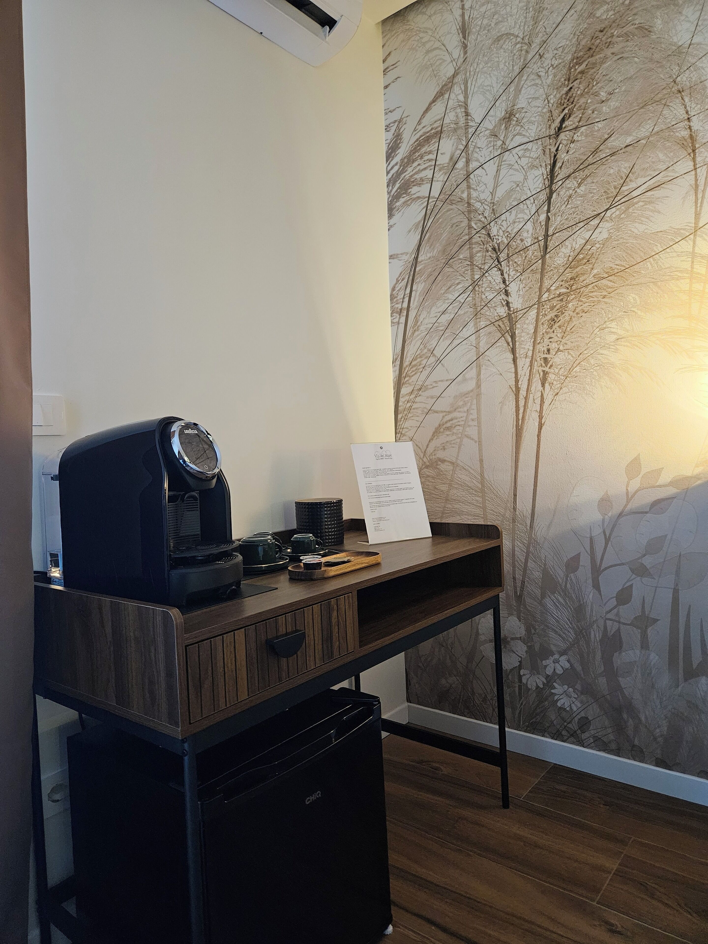 A kitchenette with a coffee machine, a kettle, and a tray, a wooden desk, and a wall with a nature-themed mural.